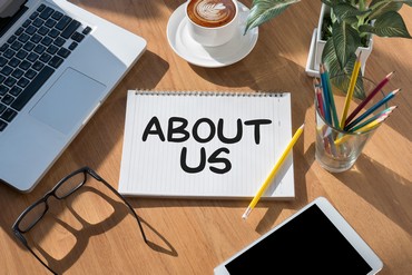 About Us printed on an open notebook in center of image, with laptop on upper left, eyeglasses laying on lower left, a cup of latte coffee at top center, a tablet at lower right, a glass with penciles, and a green plant in the upper right. All elements are on a light wood desk.